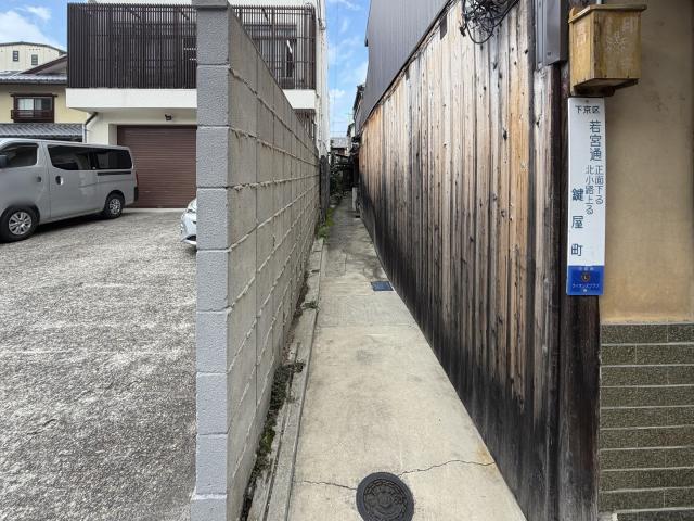 「下京区 若宮通正面下る北小路上る 鍵屋町」の町名表示板が掛かる細い路地。左にブロック塀、右に日焼けした縦板張りの外壁が奥へ続く