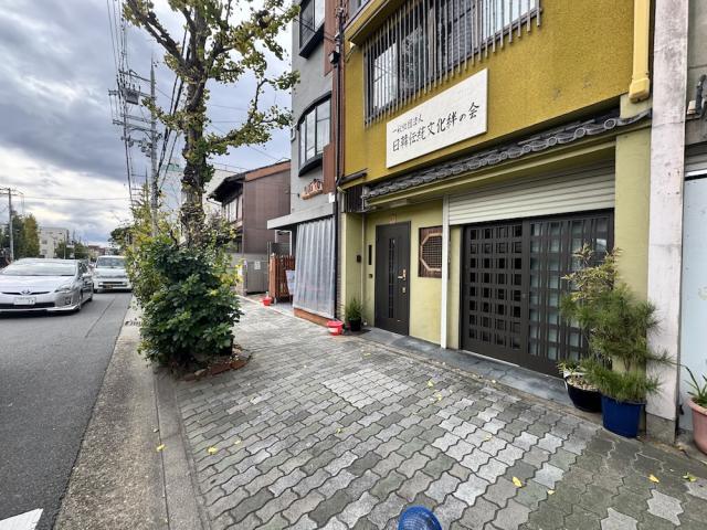 東大路通の歩道から斜めに見た1階の入口。黄色い外壁にこげ茶色の玄関ドアと格子の引き戸が並び、左手に街路樹と車道が続く