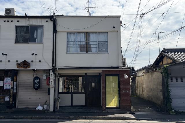 紫野雲林院町の白い外壁の建物を正面から見た外観。1階に黒い木枠の窓と木製扉、右側に赤レンガ壁と黄色いガラス戸が並ぶ
