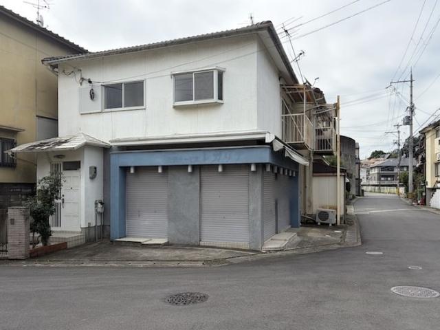西京区樫原蛸田町の店舗外観。白い2階建ての1階に青い枠とグレーのシャッター2枚、左に庇付きの玄関、右手に前面道路が延びる