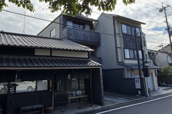 左隣の町家側から斜めに見上げた外観。瓦屋根越しに3階の窓とタイル張りのバルコニーが見え、右手に道路が続く