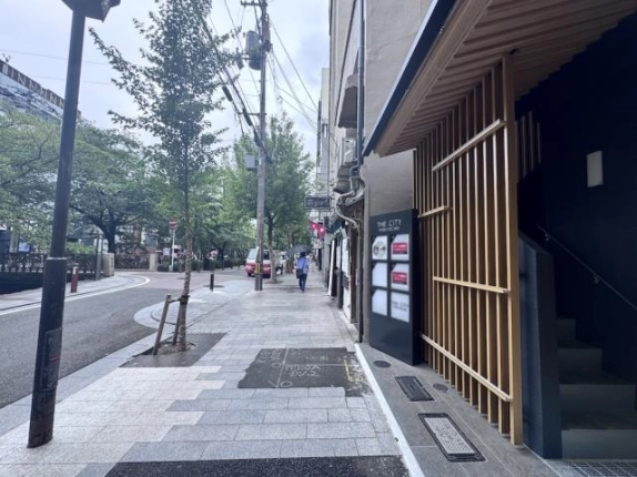 建物の脇から見た木屋町通の歩道と街路樹。右手に木製格子と館名サイン、左手に車道が続く