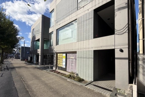 歩道沿いから見た建物の正面。2階に大きな窓、1階にポスターの貼られたガラス面と植え込みがあり、右手に奥へ続く入口通路がある