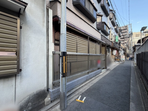 斜め前から見た建物と路地。電柱の奥に格子フェンスとひさしが続き、路地の先には張り出し窓のある建物や提灯を下げた店、自転車が見える