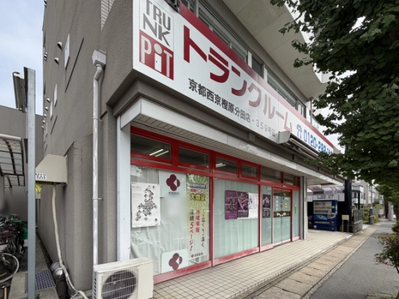 反対側の角から見たグレーの外壁の外観。大きな白い看板の下に赤い枠のガラス張り店舗、右端に自動販売機とイチョウの木