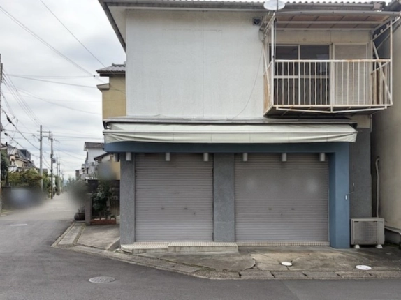 日よけテントの下にシャッター2枚が並ぶ別の面。2階右側にベランダ、右下にエアコン室外機、左手に道路が続く