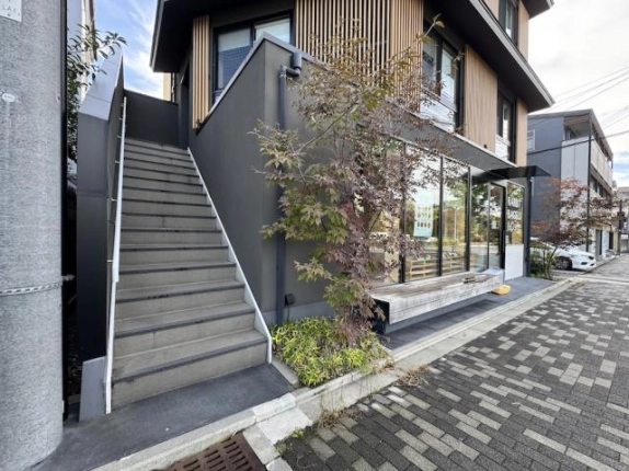 2階へ続く外階段の近景。グレーの段とダークグレーの壁の脇に、モミジの植栽と店舗前のベンチが見える