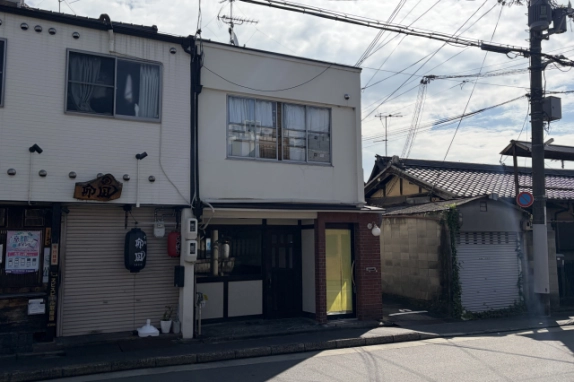 道路越しに見た1階店舗の正面。左隣に提灯を下げた店、右側に瓦屋根の家屋とシャッター、駐車禁止標識の付いた電柱が並ぶ