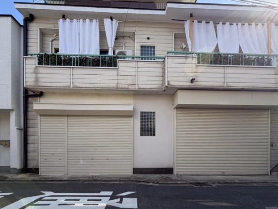 新桝屋町店舗の正面外観。1階は左に庇付きの2連シャッター、中央にガラスブロック窓、右に大きなシャッターが並び、2階に緑の手すりのバルコニーがある