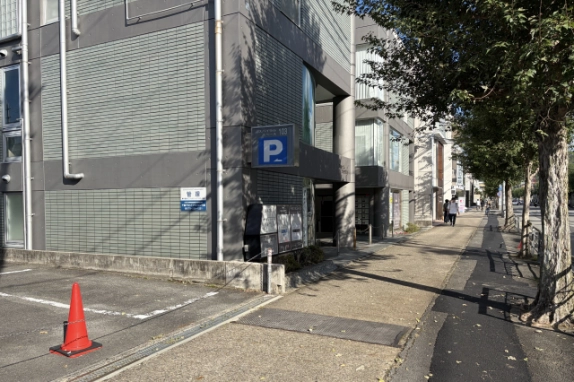 駐車場の角から見た1階入口まわり。「P」看板と「管理」表示、集合ポストが並び、右手には街路樹の続く広い歩道が延びる