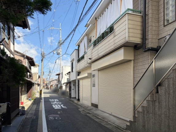 右手前から通りの奥を望む外観。手前に大きなシャッターと外階段、通りの左側には木造の町家と電柱が続く
