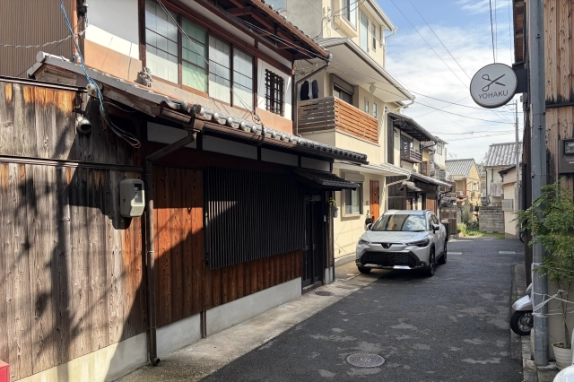 町家1階の縦格子と瓦の庇を斜めから見た様子。右手にはさみ柄の丸い看板が下がり、住宅が並ぶ路地が奥へ続く
