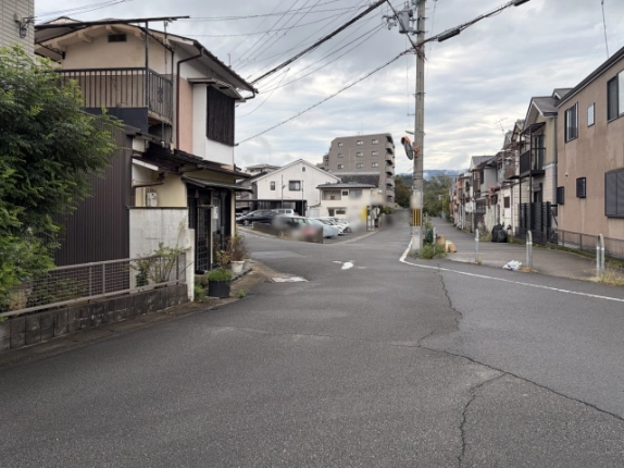 建物前の道路から見た周辺の街並み。左に店舗、奥にマンションと駐車場、遠くに山並み
