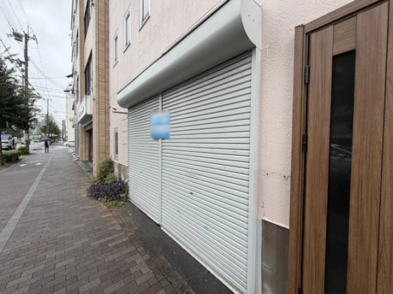 1階店舗のシャッターと出入口を斜めから見た様子。建物前にレンガ調の歩道が続く