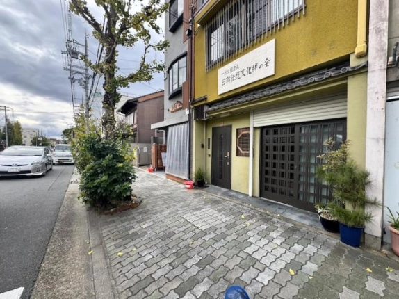 東大路通の歩道から斜めに見た1階の入口。黄色い外壁にこげ茶色の玄関ドアと格子の引き戸が並び、左手に街路樹と車道が続く