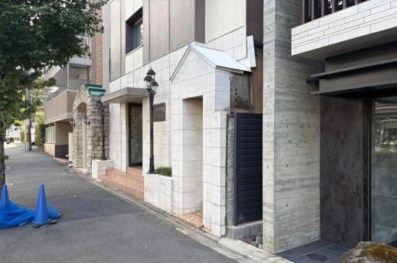 白いタイル張りの1階外壁を斜めから見た様子。切妻形の白い壁と黒いルーバー扉、街灯が並び、奥に石積みのアーチが見える