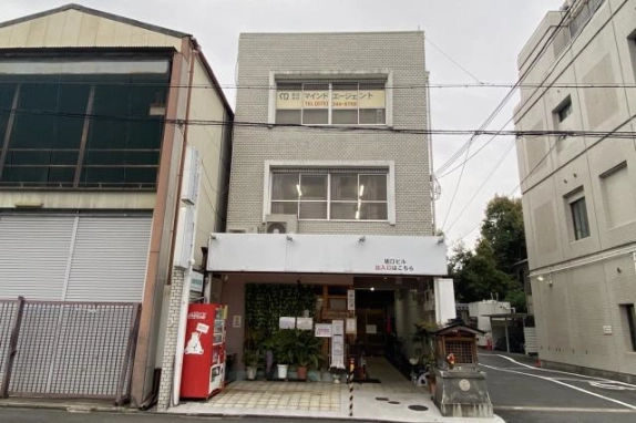 下京区の坂口ビルを正面から見た外観。白いタイル張りの3階建てで、2・3階に横長の窓、1階に「坂口ビル 出入口はこちら」の白い看板と赤い自動販売機がある
