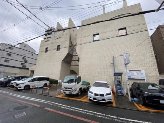 美濃利ビルを左斜め前から見た外観。白いタイル張りの外壁に尖った塔状の飾りがあり、網目状の庇がかかる入口の前に駐車区画が並ぶ