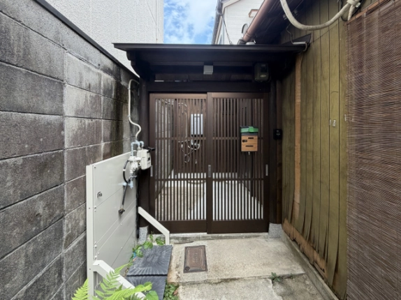 焦げ茶色の格子引き戸を正面から見た様子。戸の左にガスメーター、戸に郵便受け、右の柱にインターホンが付き、上に小屋根が架かる