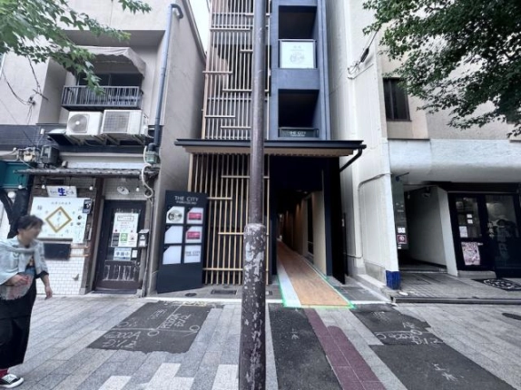 正面から見た新築ビルの低層部。左に木製格子と館名サイン、右に紺色の壁面と1階の細長い入口通路
