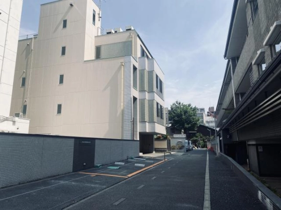 少し離れて見た側面外観。ベージュの壁に小窓が縦に並び、屋上に設備機器が見える。まっすぐ延びる道路の先には樹木と町並みが続く