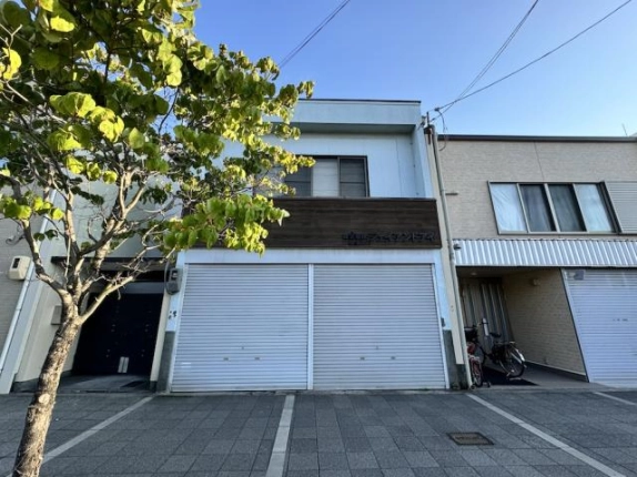 下京区花畑町の貸店舗事務所を歩道から正面に見た外観。2階に窓、中央に木目の看板帯、1階は2枚並びのシャッターで、左手前に街路樹