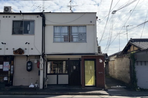 紫野雲林院町の白い外壁の建物を正面から見た外観。1階に黒い木枠の窓と木製扉、右側に赤レンガ壁と黄色いガラス戸が並ぶ