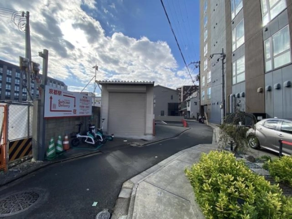 東九条東山王町の細い路地。左に「京都駅」と書かれた案内看板と電動バイク、正面にシャッター付きの平屋倉庫、右に大型建物の外壁と植栽が並ぶ