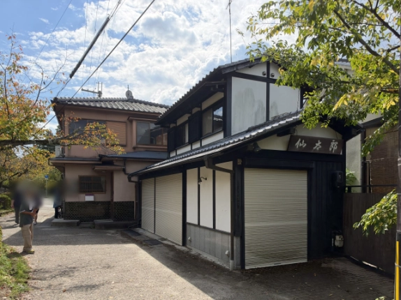 木製看板のある妻側と正面のシャッターを斜めから見た外観。左隣にピンク色の外壁の建物が建つ