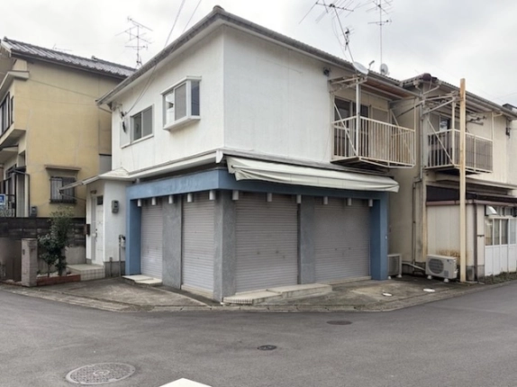 道路の角から斜めに見た2階建て店舗の全景。2つの面にシャッターが並び、右側の面にテントと2階のベランダが付く