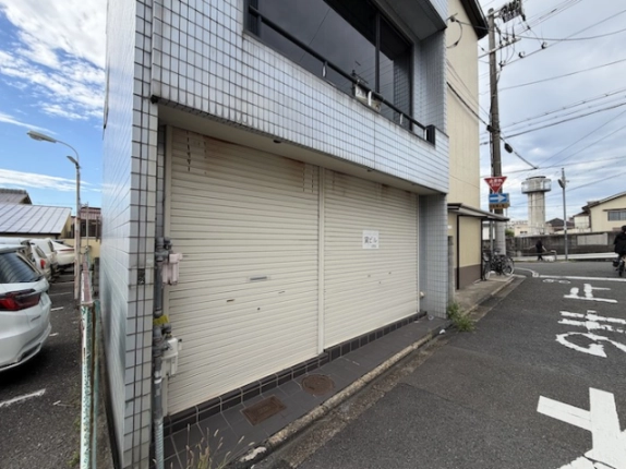 左斜め前から見た1階のシャッターと建物の角。左側に隣の駐車場、右側に「止まれ」の路面表示がある交差点