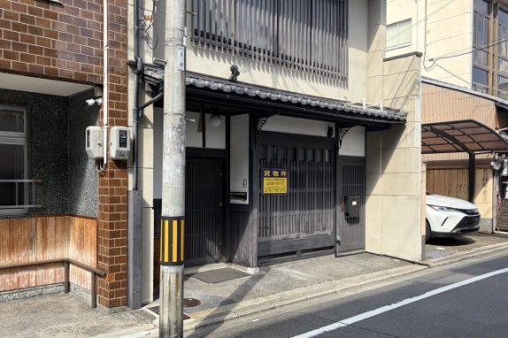 1階入口まわりの近景。瓦の庇の下に格子戸と玄関扉、黄色い貸物件の看板があり、手前に電柱が立つ