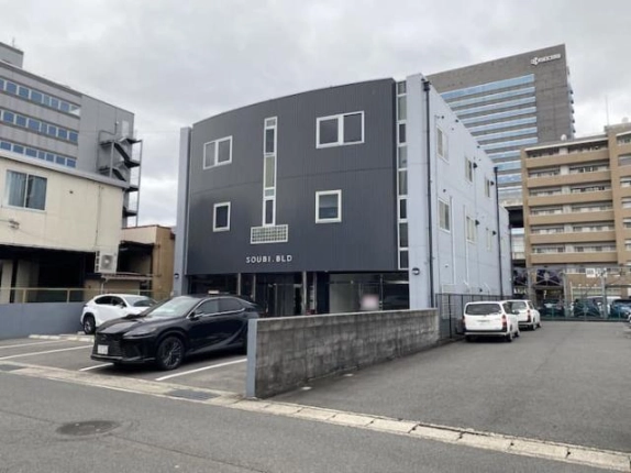 道路の向かいから見たSOUBI BLDと周辺の街並み。ブロック塀の両側に駐車場があり、建物の背後に高層ビルとマンションが建つ