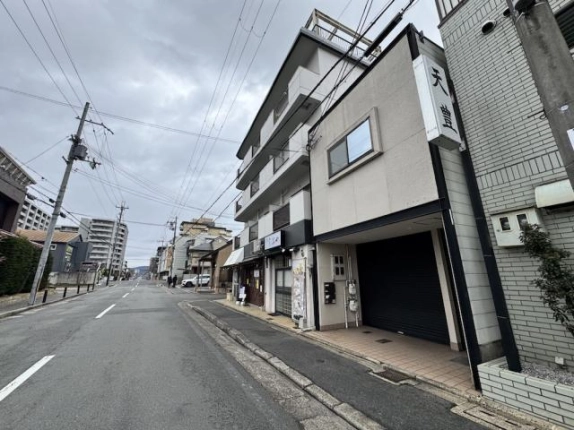 通りの左手から見上げた建物と周辺の街並み。中層マンションの右隣に建ち、1階にシャッター付きの区画がある