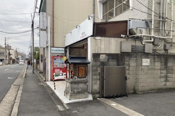 京都駅から徒歩8分の間之町通沿い、歩道から見た1階入口付近。赤い自動販売機と小さな祠、ブロック塀の門扉が並び、左手に通りが続く