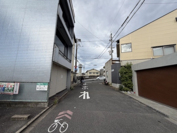 反対側から見た前面道路。左側に月極ガレージの看板がある駐車場とタイル張りの3階建、道の先に交差点がある