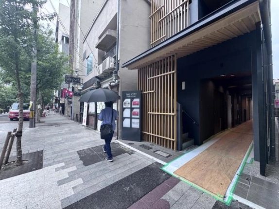 軒下から奥へ続く1階の入口通路。木製の軒天と縦格子があり、通路左手に上階への階段が見える