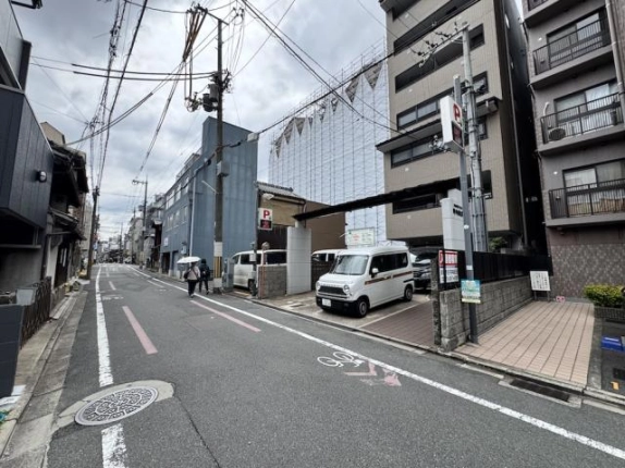 四条駅から徒歩9分の通りに面したビルと門前の駐車スペース。手前にマンホール、左奥へ続く町並みと青い外壁の建物が写る