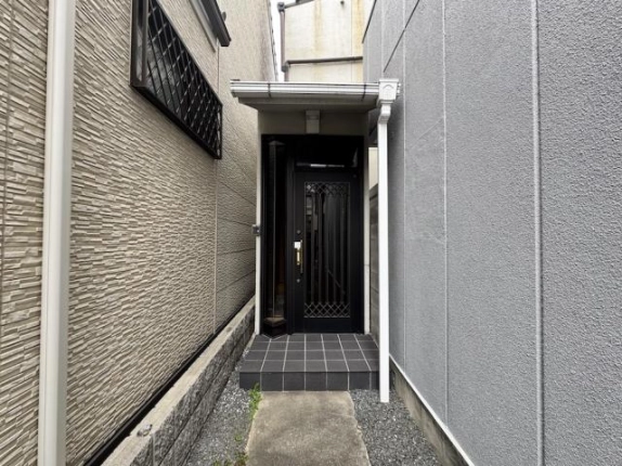正面から見た玄関までの通路。左右の外壁に挟まれたコンクリートの通路と砂利の先に、黒い玄関ドアとタイルの段差