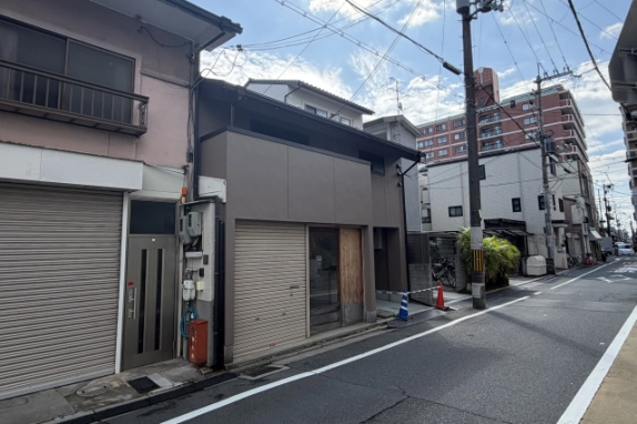 左隣のピンク色の建物側から見た斜めの外観。建物の先に道路が続き、奥にレンガ色のマンションが建つ