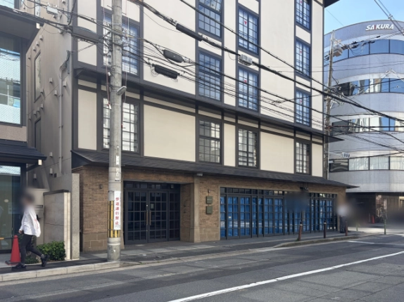 下京区の通りから斜めに見た外観。1階左に黒い格子の両開き玄関扉、右に青いシートが掛かるガラス戸の区画、上階は白壁に濃い色の格子窓が並ぶ