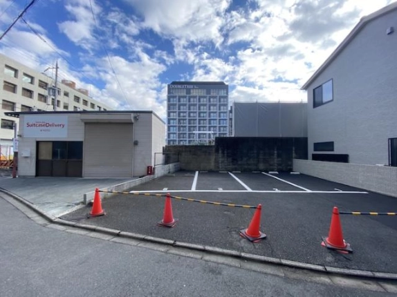 道路の正面から見た白線区画の舗装スペース。手前にカラーコーンと黄黒のバー、奥にブロック塀と高層ホテル、左にシャッター付き倉庫が建つ