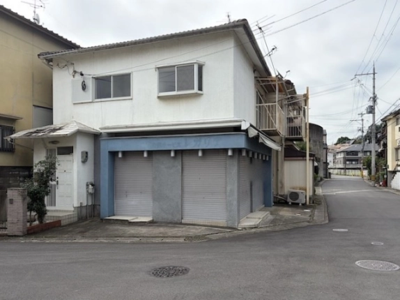 西京区樫原蛸田町の店舗外観。白い2階建ての1階に青い枠とグレーのシャッター2枚、左に庇付きの玄関、右手に前面道路が延びる