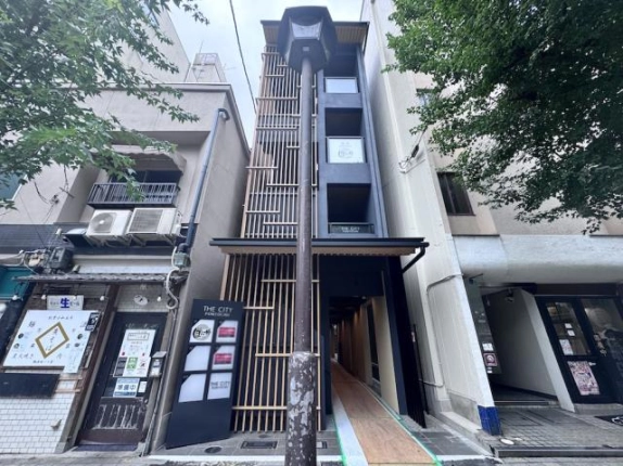 見上げて撮影した建物全景。上階まで続く木製格子と紺色の壁面に各階の開口部が並び、手前中央に街灯が立つ