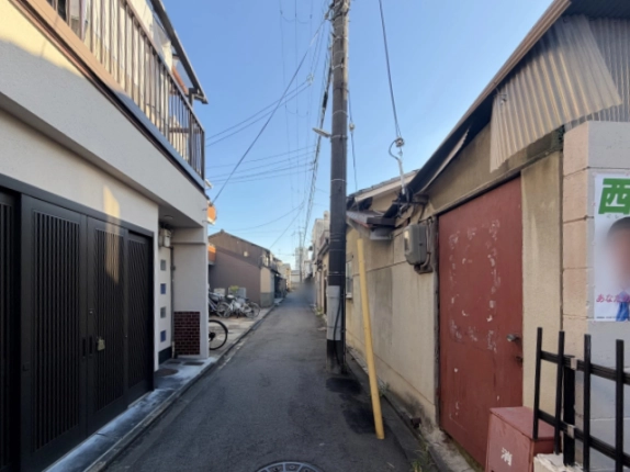 まっすぐ奥へ続く細い路地。左に黒い格子戸の住宅、右に赤い鉄扉と消火器箱