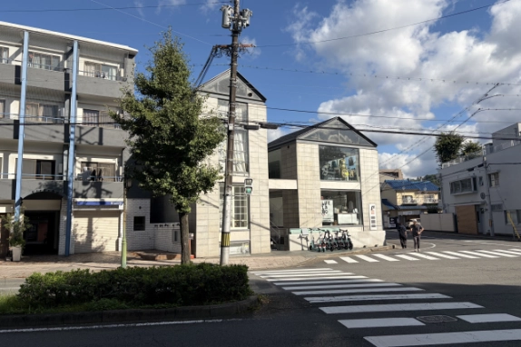 京都市北区の交差点の角に建つツリーズビル。植え込み越しに、格子窓のある三角屋根の棟とガラス張りの棟、左隣の集合住宅が見える