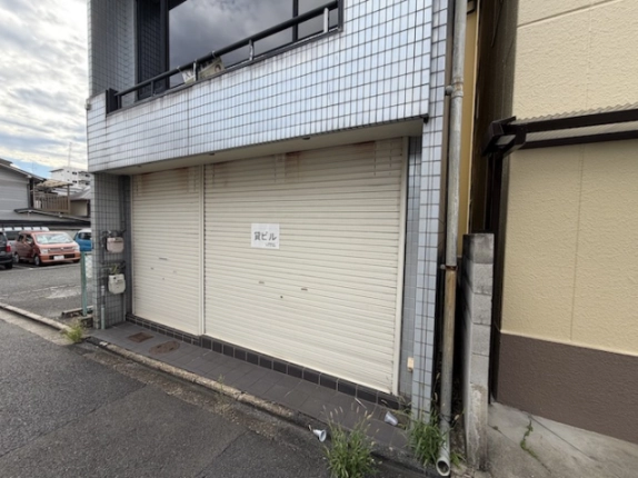 1階の店舗部分を右斜め前から見た様子。「貸ビル」の貼り紙があるシャッターとタイル張りの床、左側の壁にメーター類がある