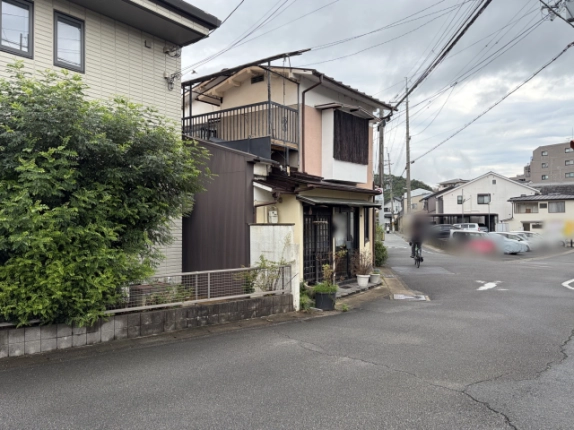 左斜め前から見た建物。手前に隣家の植栽、右側に駐車場の方へ延びる前面道路