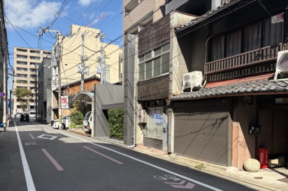 少し離れた位置から通り沿いに見た外観と周辺。右手前に隣の町家、左に自転車レーン表示のある道路が延び、奥にマンションなどが並ぶ