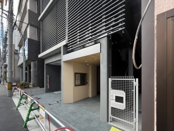 右斜めから見た1階エントランス周り。ルーバー外壁の下に集合ポストのあるベージュ壁の入口、右に白いメッシュの扉、左に屋根の下のグレーの壁の空間